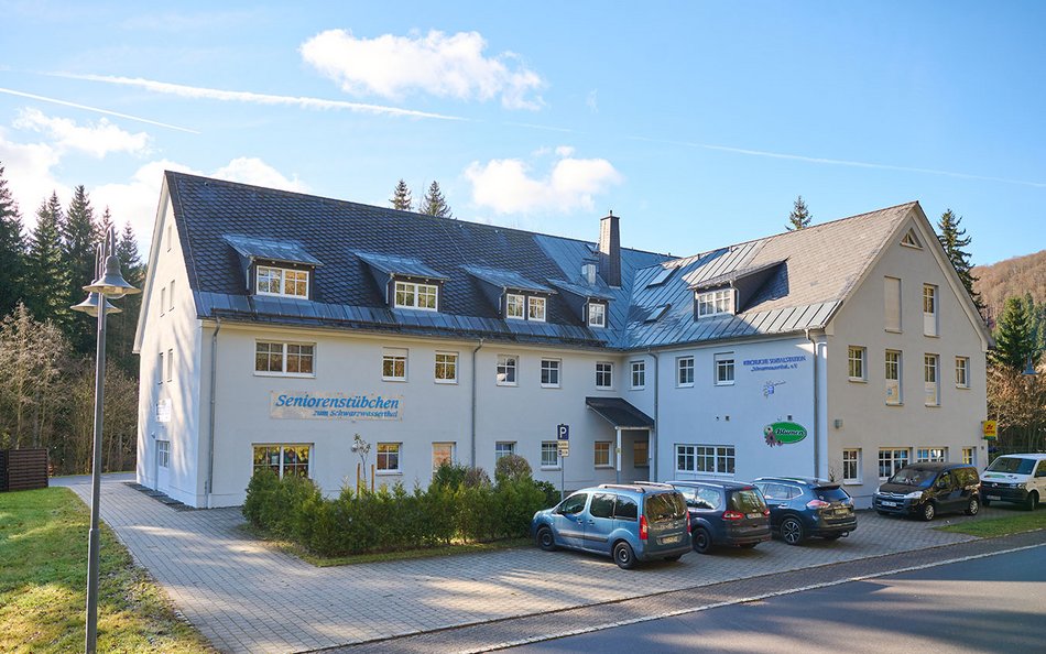 Ein großes, helles Gebäude mit schwarzem Dach und vielen Fenstern. Auf der Fassade steht „Seniorenstübchen – Haus der Betreuung und Pflege“. Mehrere Autos parken vor dem Haus auf gepflasterten Stellplätzen. Das Gebäude liegt ruhig am Waldrand, umgeben von Natur. Der Himmel ist blau mit einigen Wolken, es scheint ein sonniger Tag zu sein.