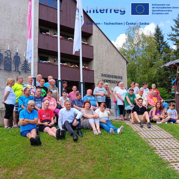 Auf dem Bild sind viele Menschen zu sehen. Sie stehen und sitzen vor einem großen Haus. Das Haus heißt: Jugendherberge Hormersdorf. Die Menschen gehören zu einer Gruppe. Es sind Frauen, Männer und Kinder dabei. Einige sitzen auf der Wiese, andere stehen dahinter. Alle schauen freundlich und wirken fröhlich. Oben links sieht man drei Flaggen. Rechts oben sind Logos von der Europäischen Union und dem Projekt „Interreg Sachsen – Tschechien“. Im Hintergrund stehen viele Bäume. Das Wetter ist schön – es scheint die Sonne.