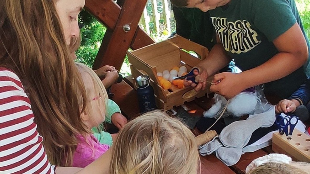 Eine Erzieherin begleitet mehrere Kinder beim gemeinsamen Basteln an einem Holztisch im Freien. Die Kinder gestalten mit Stoff, Watte und verschiedenen Materialien kreatives Spielzeug und arbeiten konzentriert zusammen. Das Bild vermittelt Ferienfreude, Kreativität und gemeinschaftliches Lernen.