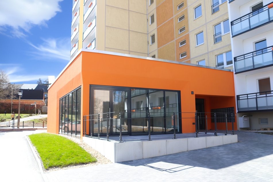 Modernes Wohngebäude mit zahlreichen Balkonen. Die Fassade ist in warmen Gelb- und Orangetönen gehalten. Vor dem Gebäude steht ein großer, grüner Baum, der einen Teil des Hauses leicht verdeckt und für eine lebendige, freundliche Atmosphäre sorgt. Der Himmel ist blau mit weißen Wolken, was dem Bild einen hellen, positiven Gesamteindruck verleiht. Die Darstellung wirkt bewusst grafisch verfremdet – wie ein digital bearbeitetes oder gemaltes Bild – und legt den Fokus auf städtisches Wohnen in einer grünen Umgebung.