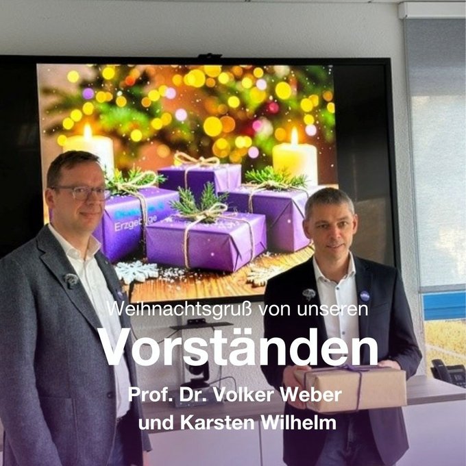 Auf dem Bild sind zwei Männer in Businesskleidung zu sehen, die nebeneinander in einem Raum stehen. Beide tragen dunkle Anzüge und helle Hemden. Einer der Männer hält ein Geschenk in den Händen, vor ihnen liegen weitere verpackte Präsente auf einem Tisch.  Im Hintergrund befindet sich ein großer Bildschirm. Darauf ist ein weihnachtliches Motiv mit Kerzen, Tannengrün, Lichtern und lilafarbenen Geschenkpaketen zu sehen. Über das Bild ist Text eingeblendet mit der Aufschrift: „Weihnachtsgruß von unseren Vorständen – Prof. Dr. Volker Weber und Karsten Wilhelm“.  Die Szene vermittelt eine festliche, wertschätzende Atmosphäre und steht im Zusammenhang mit einem offiziellen Weihnachtsgruß der Diakonie Erzgebirge.