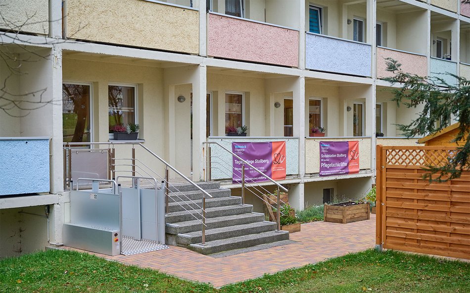 Das Bild zeigt die Außenansicht eines Wohn- oder Pflegegebäudes mit mehreren Balkonen, die in verschiedenen Pastellfarben gestrichen sind. Vor dem Eingang befinden sich eine kleine Treppe und ein barrierefreier Plattformlift, der einen ebenerdigen Zugang ermöglicht. Bunte Banner an den Geländern weisen auf soziale Angebote wie Tagespflege und die Suche nach Pflegefachkräften hin. Vor dem Gebäude sind ein kleiner Gartenbereich mit Hochbeeten und ein Sichtschutzzaun zu sehen. Die Atmosphäre wirkt freundlich und funktional, mit einem Fokus auf Zugänglichkeit und Pflegeangebote.