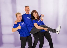 Drei Pflegekräfte in blauer Kleidung posieren fröhlich und heben Kollegin hoch