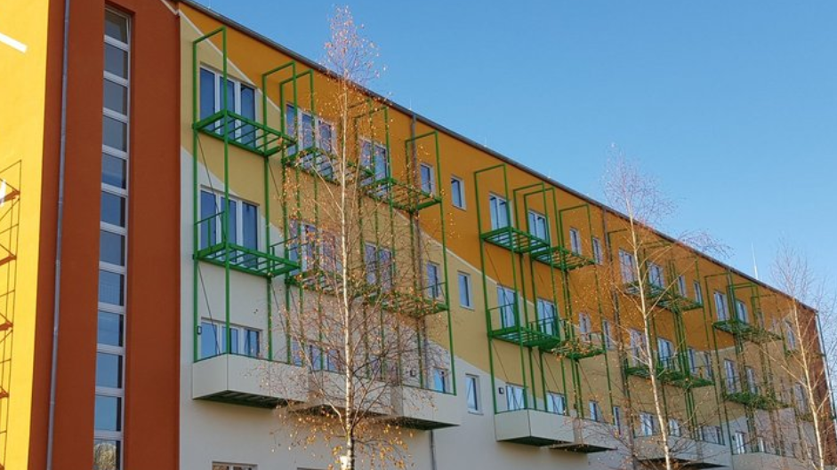 Das Foto zeigt ein modernes, farbenfrohes Gebäude mit mehreren Stockwerken. Die Fassade ist in einem lebendigen Mix aus Orange, Gelb und Beige gestaltet. Besonders auffällig sind die grünen Balkongestelle, die sich regelmäßig über die Front verteilen und dem Gebäude einen frischen, dynamischen Charakter verleihen. Schlanke Bäume mit kahlen Ästen deuten auf Herbst oder Winter hin. Der Himmel ist wolkenlos und strahlend blau – das Sonnenlicht bringt die Farben der Fassade besonders schön zur Geltung. Die Architektur wirkt offen, freundlich und einladend.