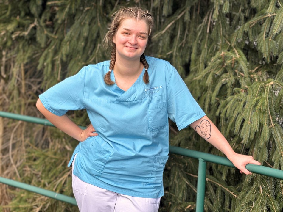 Person in blauer Pflegekleidung steht lächelnd draußen vor grünen Bäumen