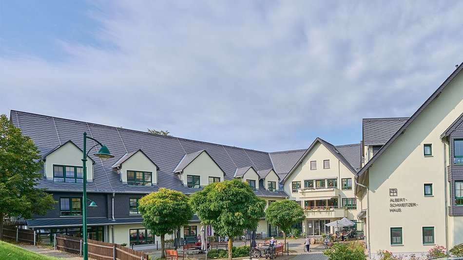 Das Bild zeigt das die Tagespflege im „Albert-Schweitzer-Haus“, eine moderne und freundlich gestaltete Pflege- oder Betreuungseinrichtung mit mehreren Etagen und Satteldächern. Die helle Fassade mit dunklen Fensterrahmen wirkt einladend und gepflegt. Vor dem Gebäude befindet sich ein begrünter Hof mit schattenspendenden Bäumen, Sitzbänken und Sonnenschirmen, der Bewohner:innen und Besucher:innen als Begegnungs- und Erholungsort dient. Menschen sind draußen aktiv – einige im Rollstuhl, andere im Gespräch – was auf eine lebendige Gemeinschaft und soziale Teilhabe hinweist. Das Bild vermittelt ein Gefühl von Geborgenheit und Lebensqualität im Alter.