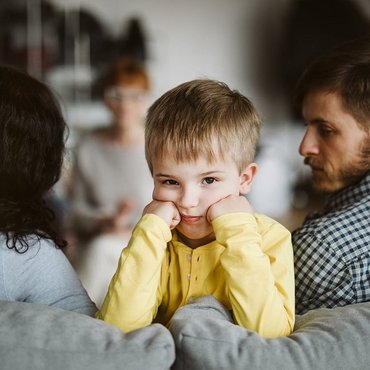 Auf dem Bild sitzt ein Kind auf einem Sofa. Das Kind trägt ein gelbes Shirt und stützt den Kopf auf die Hände. Es schaut nach vorne und sieht traurig oder nachdenklich aus. Links und rechts neben dem Kind sitzen zwei Erwachsene. Die Erwachsenen schauen sich nicht an. Sie schauen weg und sehen auch traurig oder wütend aus. Im Hintergrund sieht man eine weitere Person, die unscharf ist. Das Bild zeigt eine ernste Stimmung in der Familie.