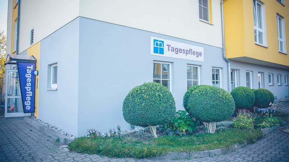 Das Foto zeigt die Außenansicht eines modernen, freundlich gestalteten Gebäudes mit gelb-grauer Fassade. Am Eingang ist ein großes, gut sichtbares Schild mit dem Wort „Tagespflege“ und dem Logo der Diakonie angebracht. Der Zugang zur Einrichtung ist über einen barrierearmen Weg möglich. Vor dem Gebäude befinden sich gepflegte, rund geschnittene Sträucher und ein kleiner Blumengarten, die eine einladende Atmosphäre schaffen. Das Bild vermittelt einen ersten positiven Eindruck von der Einrichtung und steht für professionelle Pflege in einem wohnlichen Umfeld.