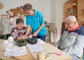 Eine betreuende Fachkraft unterstützt zwei ältere Menschen bei einer kreativen Bastelarbeit an einem Tisch. Alle wirken konzentriert und freundlich; auf dem Tisch liegen Bastelmaterialien und Werkzeuge.