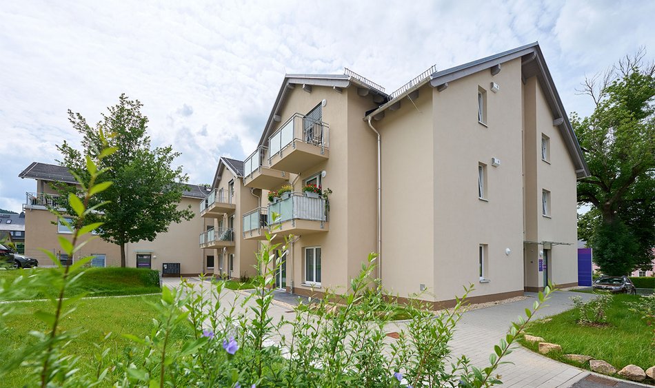 Das Bild zeigt ein modernes, helles Wohngebäude mit mehreren Balkonen, auf denen teilweise Blumenkästen zu sehen sind. Die Fassade ist in einem freundlichen Beigeton gehalten, die Architektur wirkt offen und einladend. Die gepflegte Außenanlage mit grüner Wiese, Pflanzen und einem großen Baum vermittelt Ruhe und Wohnqualität. Die barrierearme Bauweise lässt darauf schließen, dass das Gebäude möglicherweise für betreutes Wohnen oder altersgerechtes Wohnen konzipiert ist. Die Szenerie strahlt Sicherheit, Geborgenheit und ein angenehmes Wohnumfeld aus.