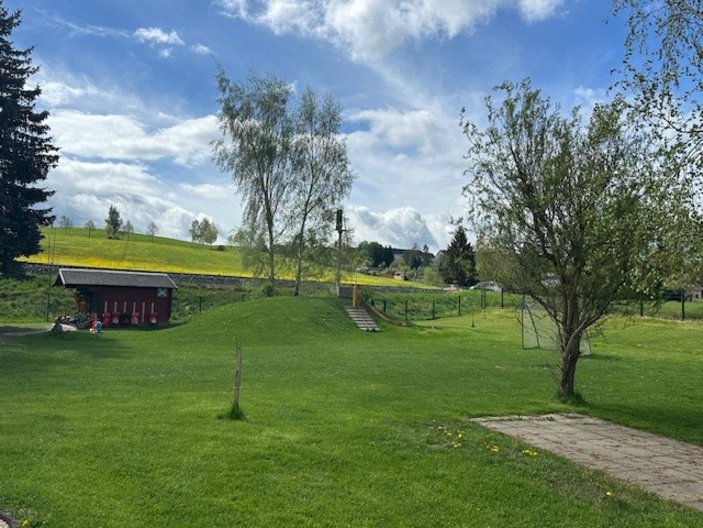 Grünes Außengelände mit Rasen, Bäumen, Gartenhaus, gepflastertem Weg und hügeliger Landschaft im Hintergrund.