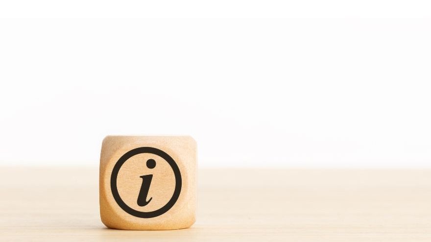 Das Bild zeigt einen Holzwürfel, der auf einer hellen Holzfläche steht. Auf der Vorderseite des Würfels ist ein schwarzes Informationssymbol („i“) in einem Kreis zu sehen. Der Hintergrund ist hell und unscharf, wodurch der Würfel deutlich im Vordergrund steht.