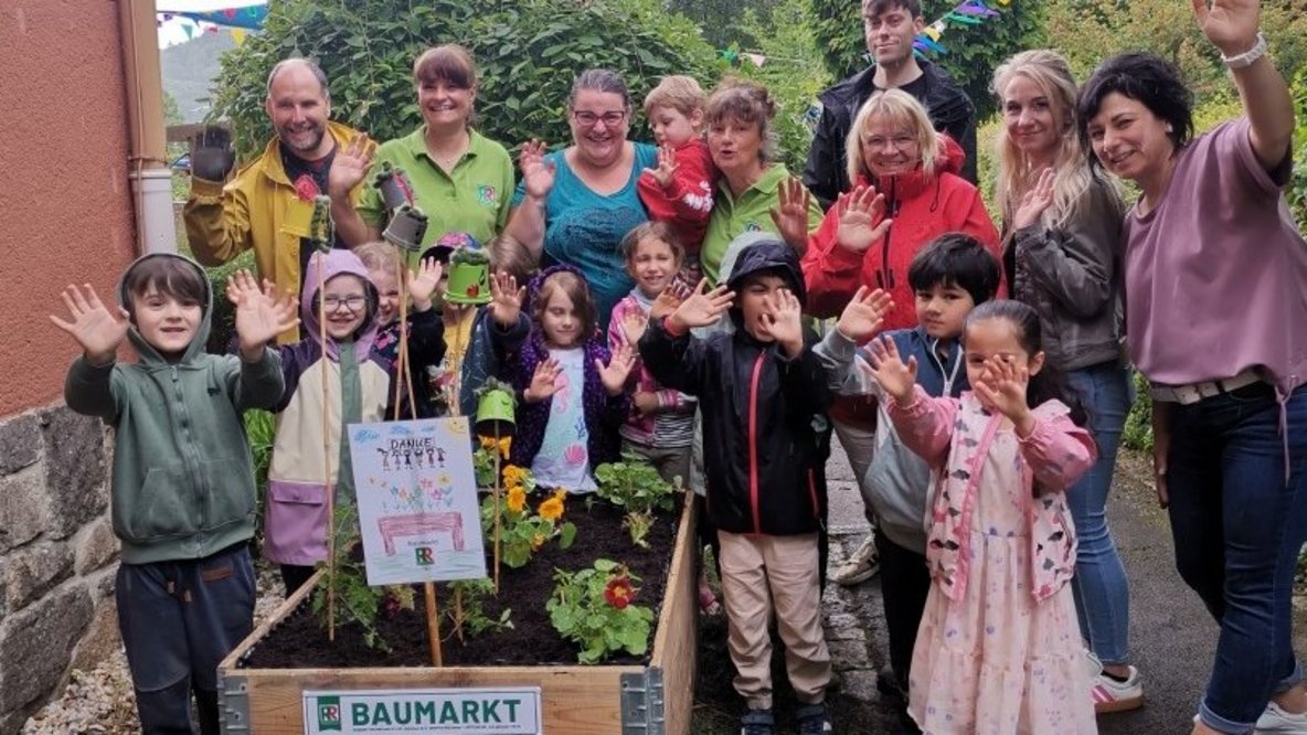 Geschafft: Gemeinsam präsentieren die Kinder, Mitarbeitenden der Kindertagesstätte, die engagierte Mutter aus dem Elternrat sowie die Unterstützer des Baumarkts Roth das fertig bepflanzte Hochbeet. Mit Blumen, Kräutern, Salaten und Gemüsepflanzen ist ein neuer Lern- und Erlebnisort entstanden, an dem die Kinder künftig säen, pflegen, beobachten und ernten können. Das Projekt zeigt, wie durch das Zusammenwirken von Eltern, regionalen Partnern und Kita Gemeinschaft erlebbar wird und nachhaltige Erfahrungen für die Kinder entstehen.