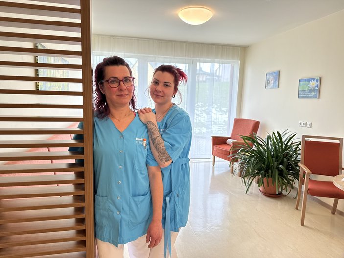 Zwei Pflegekräfte in blauer Kleidung lächeln in freundlicher Einrichtung