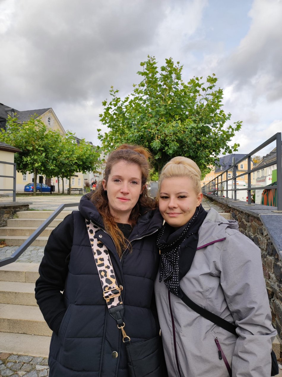 Zwei Frauen stehen draußen nebeneinander und lächeln in die Kamera