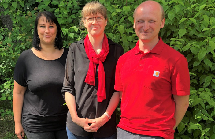 Drei Mitarbeitende stehen lächelnd nebeneinander im Freien vor grünen Büschen